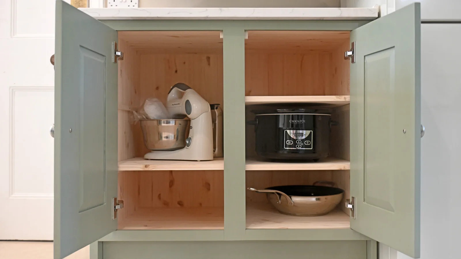 Bespoke sage green painted cupboard doors open to show pine shelving with kitchen appliances