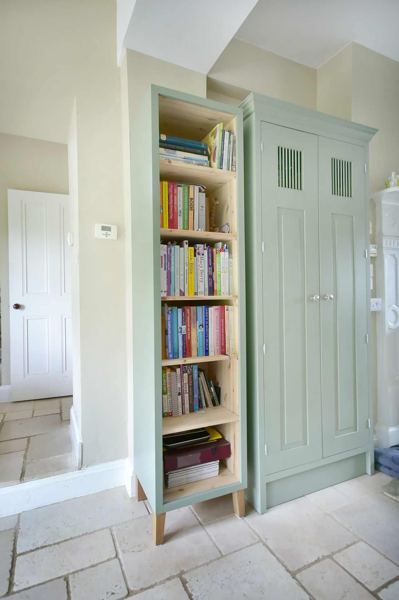 Bespoke open bookcase filled with books beside the green tall cupboard in the Malmesbury kitchen