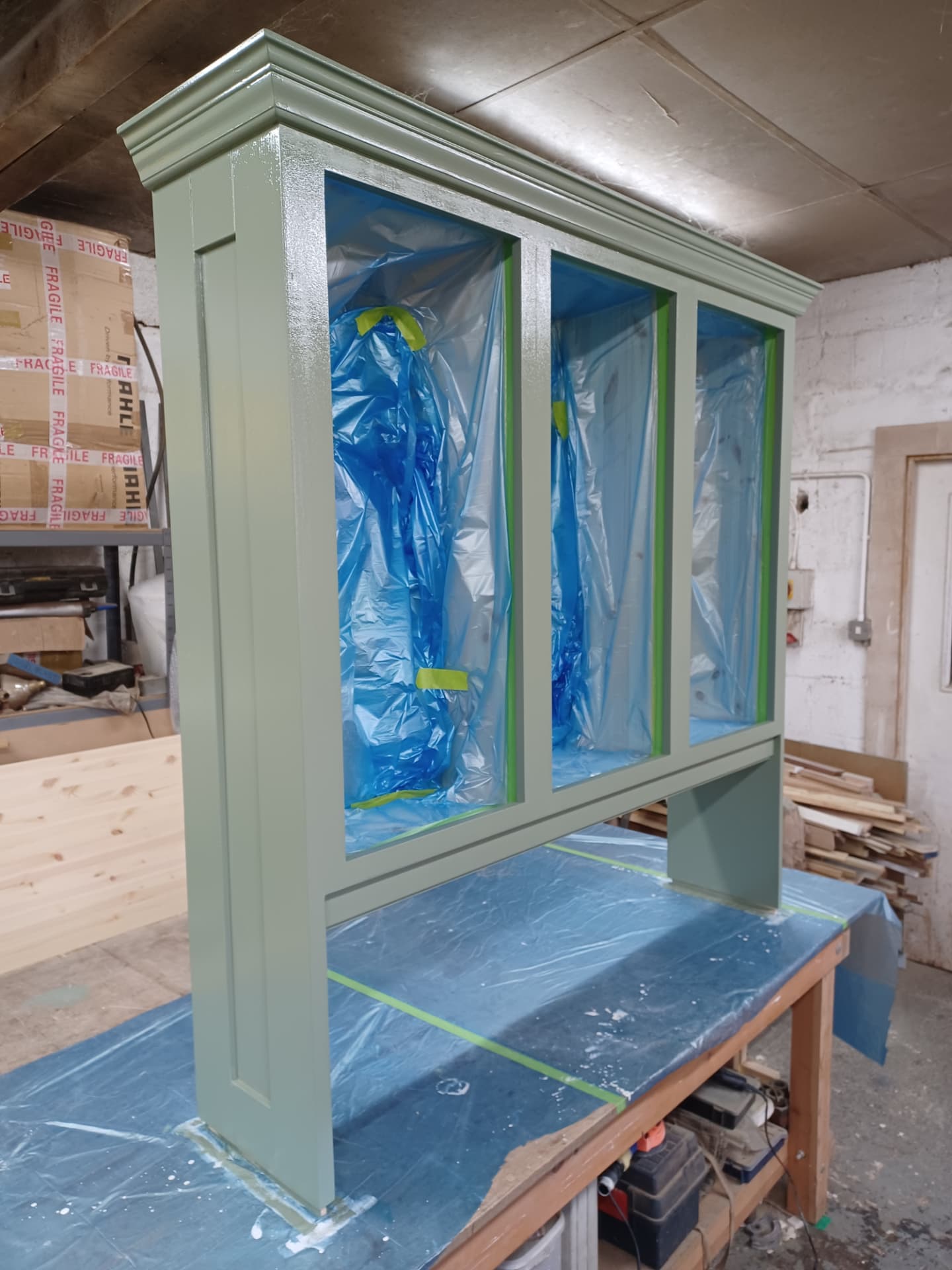 Green bespoke cabinetry with upper mouldings and masked openings during spray finishing in the workshop