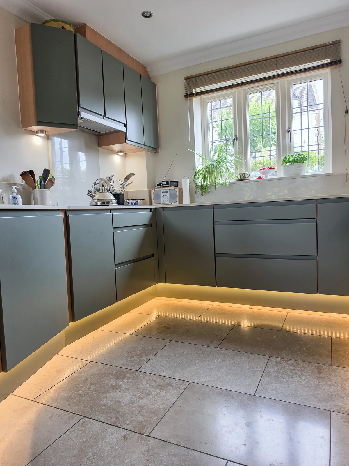 Plinth and under-cabinet LED lighting switched on beneath mid-green handleless base cabinets in the refreshed Brinkworth kitchen, reflected in the tiled floor