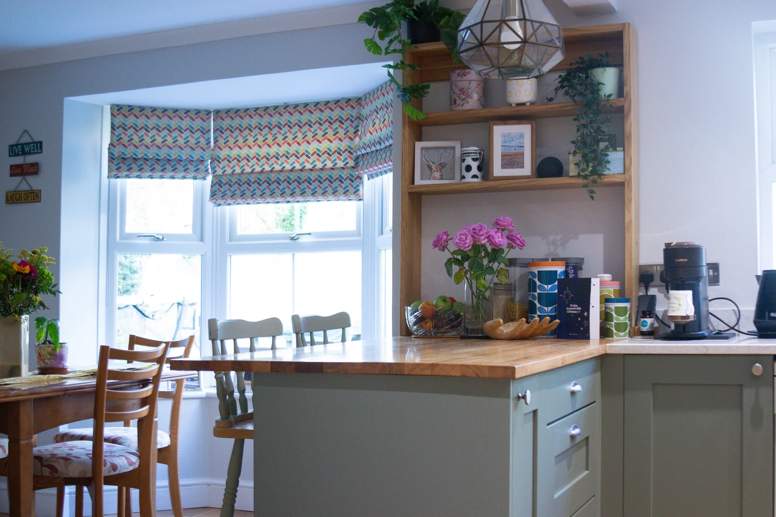 The solid oak peninsula worktop with the workshop-made oak shelving unit at its end in the Corsham custom kitchen, Regiment Green shaker cabinets alongside and the dining area beyond