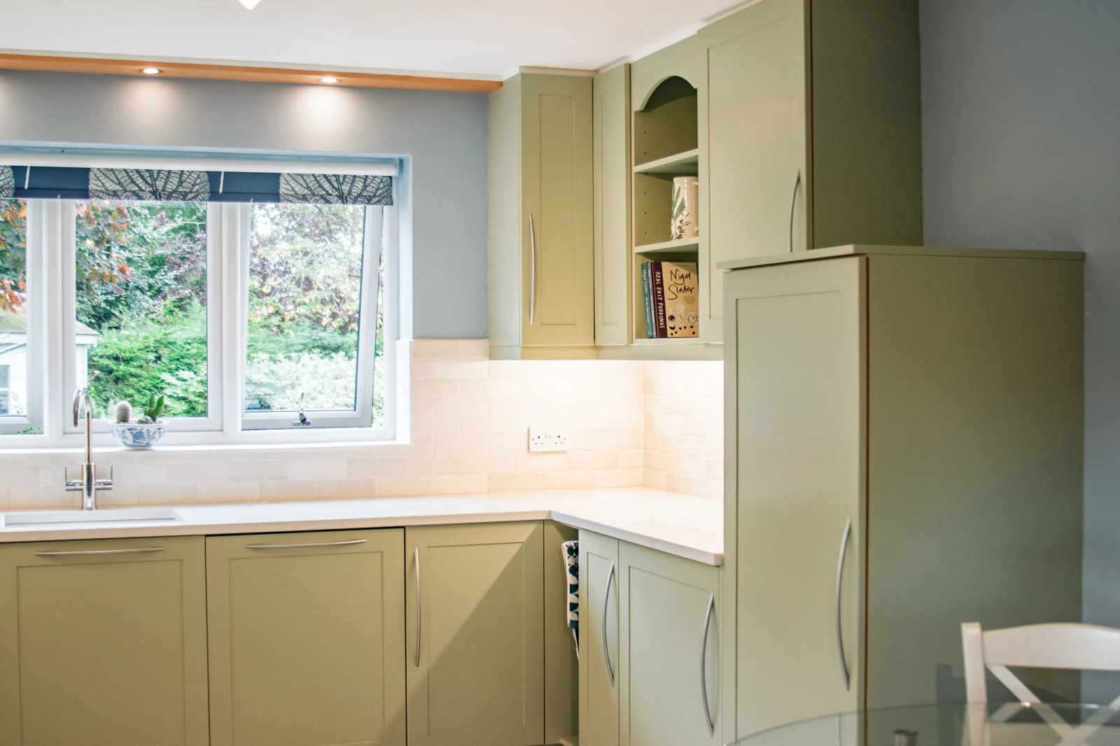 Corner run by the garden window in the Highworth kitchen, with Farrow & Ball Vert de Terre cabinets, a tall cabinet and open display shelves above lightly-veined quartz worktops