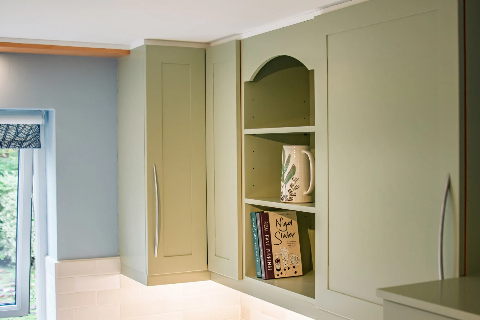 Farrow & Ball Vert de Terre wall cabinets above the corner in the Highworth kitchen, with an arched open shelf between them displaying cookbooks