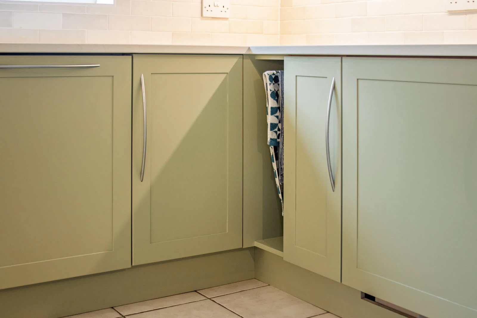 Corner base units in the Highworth kitchen, with an open tea towel cubby between the Farrow & Ball Vert de Terre cabinets and a lightly-veined quartz worktop above