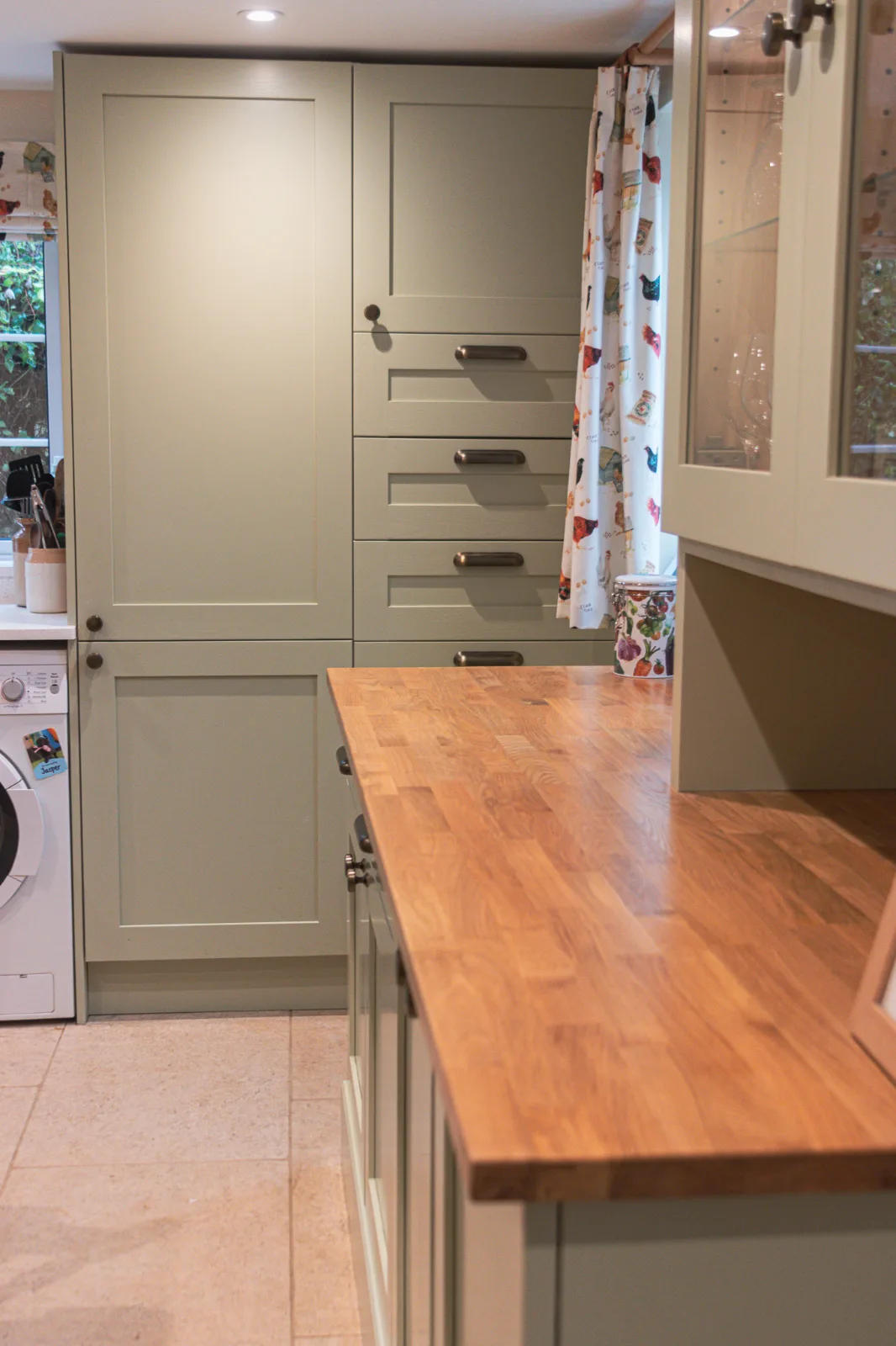 A painted kitchen larder with drawers beside a timber worktop and window.
