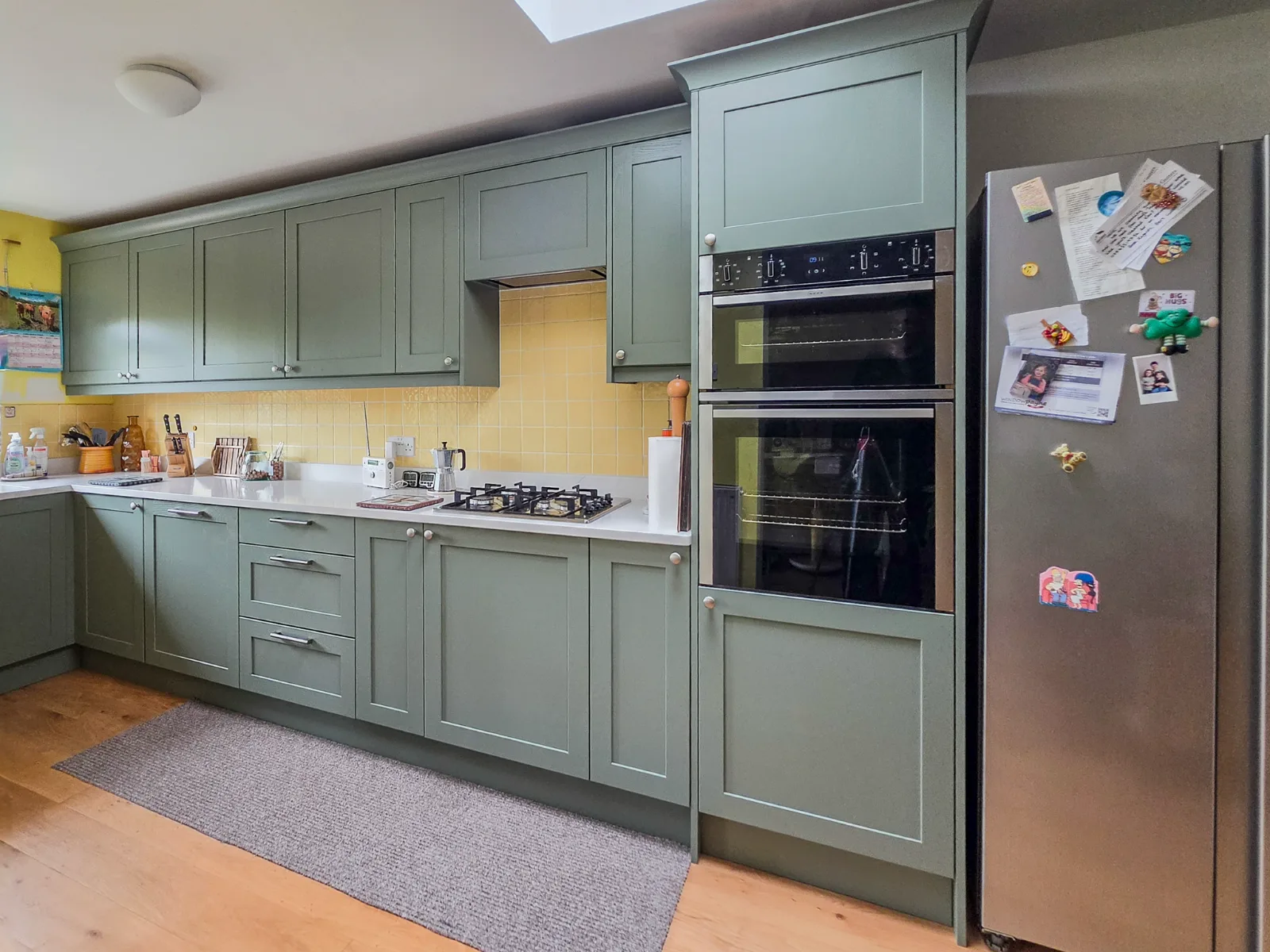 Green Shaker cabinets with pale worktops, a gas hob, stacked ovens and a freestanding fridge-freezer.
