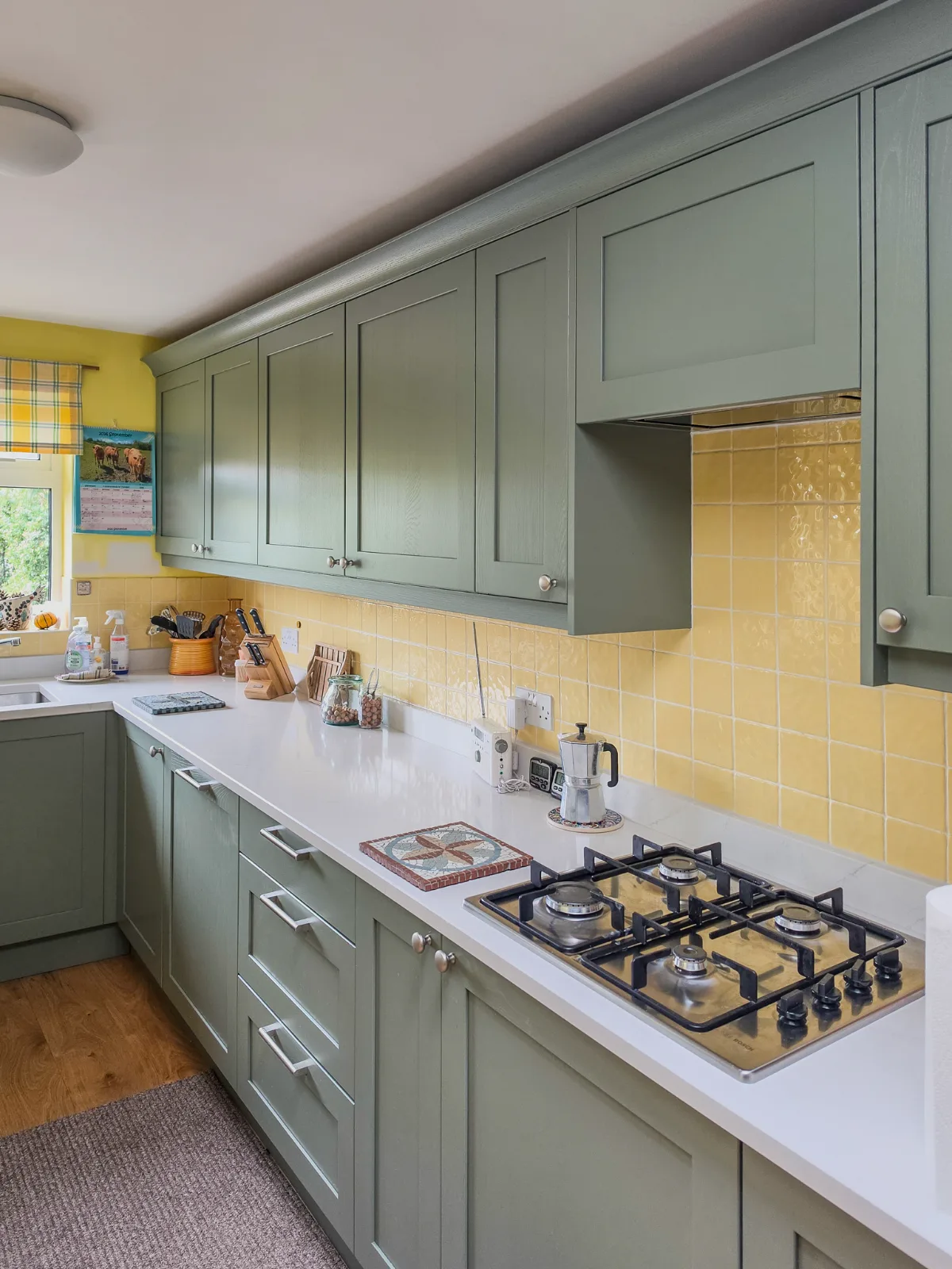 The refreshed Corsham kitchen with new sage green replacement shaker doors, a white quartz worktop and gas hob, keeping the original yellow walls and tiles.