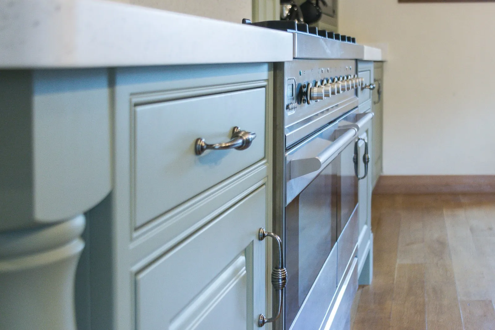 Close view of the green-painted base unit beside the stainless range cooker, with one drawer over a cupboard door, both with brushed metal handles, a light worktop above and a turned decorative column at the corner