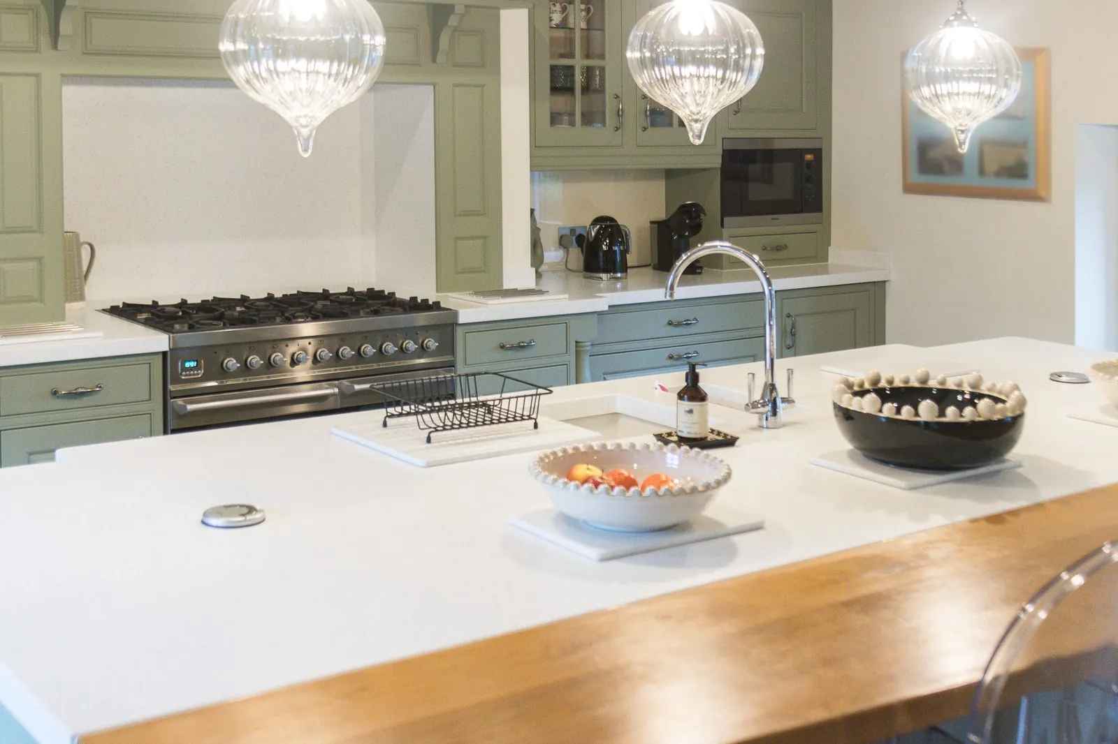 Looking across the island towards the stainless range cooker, with a light worktop, inset sink and tall curved tap on the island, a timber breakfast bar along the near edge, and beyond it the green-painted cabinetry, glazed cupboard and range-cooker mantel under three glass pendant lights