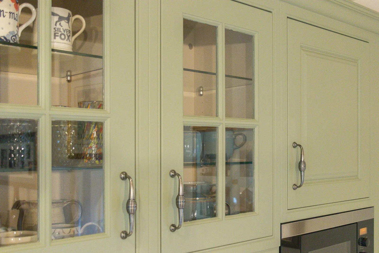 The glazed wall cabinets lit from within, with green-painted glass-fronted doors in a four-pane mullion design, cream-lined interiors and glass shelves holding mugs, glassware and blue ceramics, and the top of a built-in microwave to the right