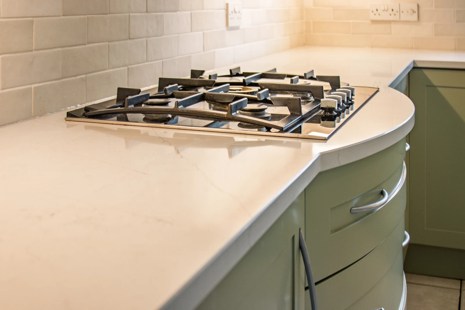 Lightly-veined white quartz worktop wrapping around curved sage-green drawers beneath the retained gas hob in the Highworth kitchen